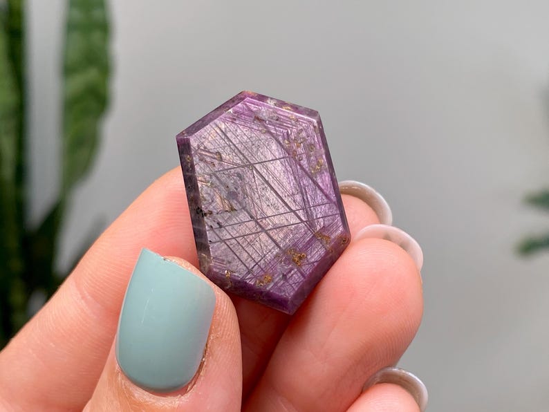 May include: A hexagonal gemstone with a deep purple colour. The stone has intricate, linear inclusions in gold and silver tones, creating a unique pattern. The gemstone is held between fingers.