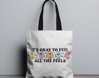 Könnte beinhalten: Weiße Einkaufstasche mit schwarzen Henkeln und dem Text "It's okay to feel all the feels" in Schwarz. Der Text ist von einer Reihe von fünf bunten Comic-Gesichtern umgeben.