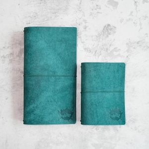 May include: Two teal leather-bound journals with a fish design embossed on the front cover. The larger journal is on the left and the smaller journal is on the right.