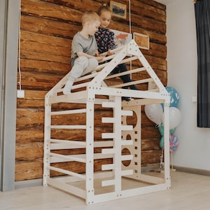 Könnte beinhalten: Ein weißer Kletterrahmen aus Holz in Form eines Hauses mit mehreren Sprossen und Aussparungen. Zwei Kinder spielen auf dem Rahmen. Der Hintergrund zeigt eine Holzwand und Luftballons.