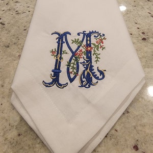 K&ouml;nnte beinhalten: Wei&szlig;e Leinen-Serviette mit einem gestickten Buchstaben M in Blau und Gold mit floralen Akzenten.