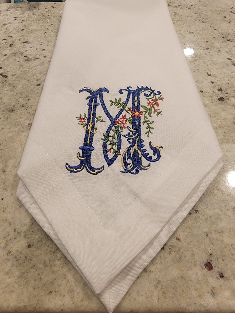 K&ouml;nnte beinhalten: Wei&szlig;e Leinen-Serviette mit einem gestickten Buchstaben M in Blau und Gold mit floralen Akzenten.