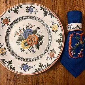 K&ouml;nnte beinhalten: Ein wei&szlig;er Keramikteller mit einem gr&uuml;nen und braunen Blumendesign. Der Teller hat einen erh&ouml;hten Rand und eine zentrale Vertiefung. Ein blauer Serviettenring mit einem wei&szlig;en Blumendesign befindet sich auf einer blauen Serviette.