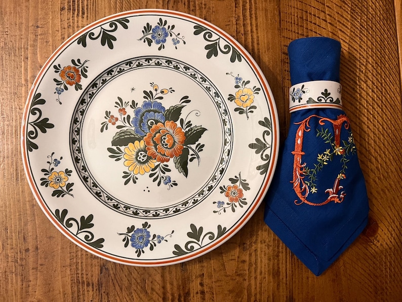 K&ouml;nnte beinhalten: Ein wei&szlig;er Keramikteller mit einem gr&uuml;nen und braunen Blumendesign. Der Teller hat einen erh&ouml;hten Rand und eine zentrale Vertiefung. Ein blauer Serviettenring mit einem wei&szlig;en Blumendesign befindet sich auf einer blauen Serviette.