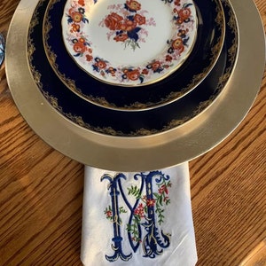 K&ouml;nnte beinhalten: Ein Satz aus drei Tellern mit goldfarbenem Rand und einem floralen Muster in Rot-, Blau- und Wei&szlig;t&ouml;nen. Die Teller sind &uuml;bereinander gestapelt und stehen auf einem Holztisch. Eine wei&szlig;e Leinenserviette mit einem blauen und roten gestickten Initial "M" befindet sich im Vordergrund.