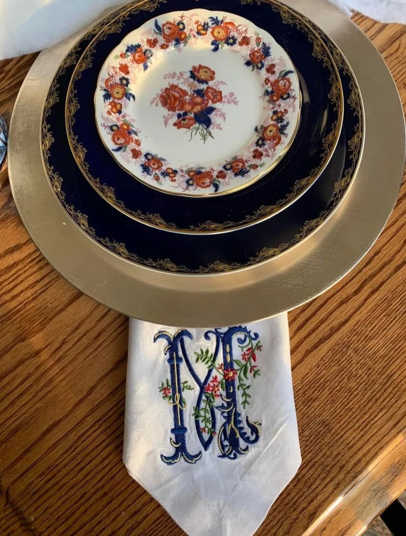 K&ouml;nnte beinhalten: Ein Satz aus drei Tellern mit goldfarbenem Rand und einem floralen Muster in Rot-, Blau- und Wei&szlig;t&ouml;nen. Die Teller sind &uuml;bereinander gestapelt und stehen auf einem Holztisch. Eine wei&szlig;e Leinenserviette mit einem blauen und roten gestickten Initial "M" befindet sich im Vordergrund.