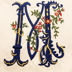 K&ouml;nnte beinhalten: Ein gestickter Buchstabe M in Blau und Gold mit floralen Akzenten. Der Buchstabe ist von roten und gr&uuml;nen Blumen und Ranken umgeben.