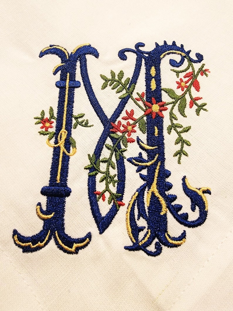 K&ouml;nnte beinhalten: Ein gestickter Buchstabe M in Blau und Gold mit floralen Akzenten. Der Buchstabe ist von roten und gr&uuml;nen Blumen und Ranken umgeben.