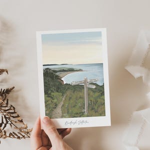 May include: A rectangular art print featuring a coastal landscape illustration with a wooden signpost in the foreground. The scene depicts a path, green vegetation, the ocean, and a pale sky. The text Budleigh Salterton is printed at the bottom.