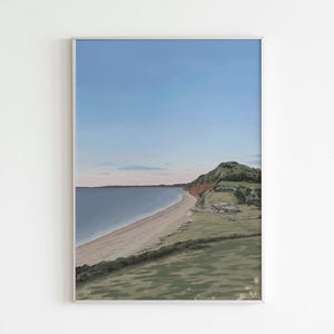 May include: A framed print of a coastal landscape. The image shows a sandy beach, a calm sea, and a green hillside under a clear blue sky. The print is enclosed in a silver frame, enhancing the artwork's presentation.