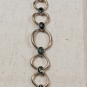 May include: A metallic chain bracelet featuring a series of alternating smooth, rounded oval links and small, dark, mottled green cabochon stones set in metal bezels. The design creates a repeating pattern of metal loops and polished stone accents.