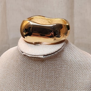 May include: A gold-colored, wide cuff bracelet with a wavy design. The bracelet is highly reflective, showing the surrounding environment. The jewelry is displayed on a neutral-toned mannequin.