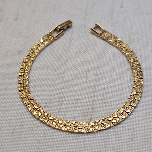 May include: A gold-toned bracelet featuring a textured, nugget-like surface pattern across the entire band. The piece is constructed with a series of interconnected links and includes a secure, hinged box clasp mechanism at the top for closure.