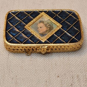 May include: A rectangular, gold-toned trinket box with a quilted navy blue lid. The lid features a central diamond-shaped portrait of a woman. The box has ornate detailing and a clasp closure, showcasing a vintage aesthetic.
