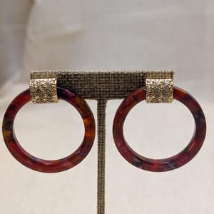 May include: A pair of circular hoop earrings featuring a marbled dark red and brown resin finish. Each hoop is attached to a gold-toned metal connector piece that is encrusted with small, clear, sparkling crystals, creating a contrast in texture.