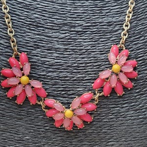 Pode incluir: Um colar dourado com três pingentes de flores. Cada flor tem pétalas rosa e rosa claro com um centro amarelo. O colar é exibido em um fundo preto texturizado.