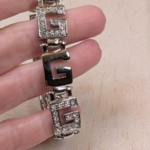 May include: A silver-toned link bracelet featuring alternating square-shaped charms. Some links are solid polished silver, while others are encrusted with clear, sparkling rhinestones. Each link is shaped like the capital letter G.