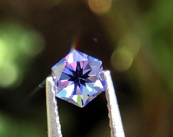 May include: A faceted, hexagonal gemstone with a deep blue and purple hue, held by silver tweezers. The gem's intricate cuts reflect light, creating a sparkling effect. The background is a soft, blurred green.