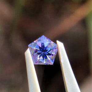 Könnte beinhalten: Ein facettierter, sternförmiger Edelstein, der von einer Pinzette gehalten wird. Der Edelstein hat eine Oberfläche mit vielen Facetten, die das Licht reflektiert und Blau- und Lilatöne zeigt. Die Pinzette ist weiß, der Hintergrund ist unscharf.
