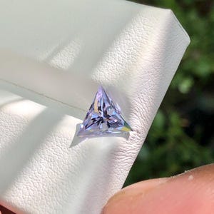 以下が含まれることがあります： 薄紫色のファセット加工された三角形の宝石。白いテクスチャ加工された表面に置かれ、背景はぼやけた緑色。宝石のファセットは光を反射し、輝きを生み出しています。