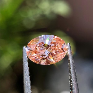 May include: A faceted, oval-cut gemstone with a peach hue is held by a pair of silver tweezers. The gem reflects light, showcasing its many facets. The background is blurred, with green and brown tones.