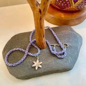 Könnte beinhalten: Eine Lavendel-Perlenkette mit einem silbernen Blumenanhänger. Die Kette liegt auf einer grauen Steinoberfläche.