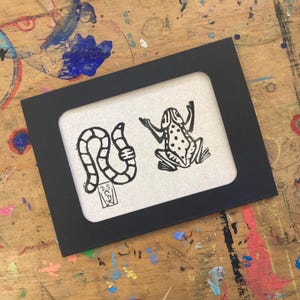 May include: A black framed print featuring two black and white illustrations. One is a stylized worm, the other a frog with spots. The print is on a textured, off-white background. The frame is rectangular with a white border.