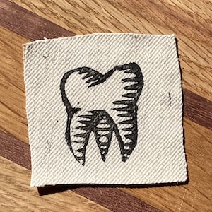 May include: A square, off-white fabric patch featuring a black, stylized illustration of a tooth. The tooth design is detailed with lines to represent the enamel and roots. The patch is likely for sewing or ironing onto clothing or accessories.