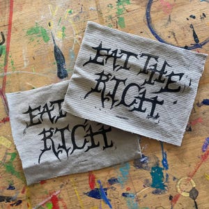 May include: Two gray fabric patches with black lettering that reads "Eat the Rich".
