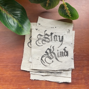 May include: A stack of four beige fabric patches with black ink lettering that reads "Stay Kind".