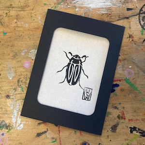 May include: A black framed print featuring a stylized black beetle illustration on a cream-colored background. The print has a rectangular opening and a small stamp in the lower right corner. The frame is black and the background is a wooden surface.