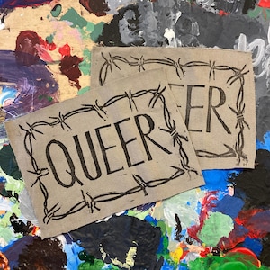 May include: Two beige fabric patches with black barbed wire borders. The top patch says "QUEER" and the bottom patch says "ER".