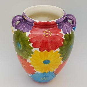 May include: A hand-painted ceramic vase with a white interior and two purple handles. The vase is decorated with large, colourful flowers in red, yellow, green, and blue, creating a vibrant floral design. The vase is a decorative item.