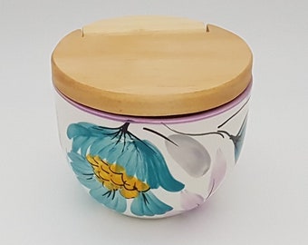 May include: A white ceramic bowl with a floral design in shades of blue, yellow, and pink. The bowl has a wooden lid with a small handle.