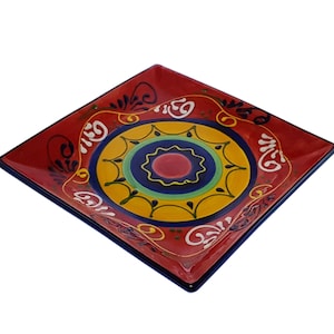 May include: Square ceramic serving dish with a red background and a colourful circular design in the centre. The design features yellow, blue, green, and red colours. The dish has a blue rim.