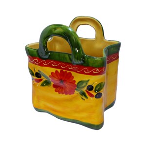 May include: A yellow ceramic utensil holder shaped like a shopping bag with a green handle. The bag has a red and green floral design with black olives.
