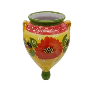 May include: A hand-painted, yellow ceramic wall vase with a red floral design. The vase has a green rim and a green base. It features two small handles and is approximately 18 cm tall.