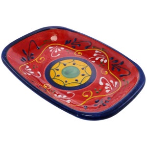 May include: A red rectangular ceramic dish with a blue rim and a colourful floral design. The centre of the dish features a yellow circle with a green centre and a white floral pattern.
