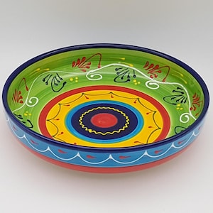 May include: A brightly coloured ceramic bowl with a hand-painted design. The bowl has a green interior with red and white floral accents, and concentric circles of red, yellow, and blue. The rim is blue with white wave patterns. A decorative kitchen item.