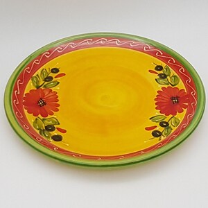 May include: A yellow ceramic plate with a green and red rim. The plate is decorated with red flowers, green leaves, and black olives.