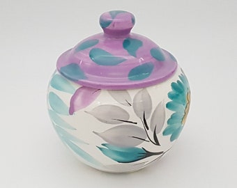 May include: A round, white ceramic jar with a lid. The lid is lavender with teal and purple accents. The jar is decorated with teal, lavender, and gray floral designs. The jar has a small, round handle on top of the lid.