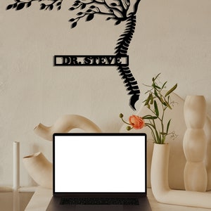 May include: A black metal wall art sculpture of a tree growing from a spine with the text "DR. STEVE" on a white wall. A laptop computer is on a white table in front of the wall art. There are white ceramic vases and flowers on the table.