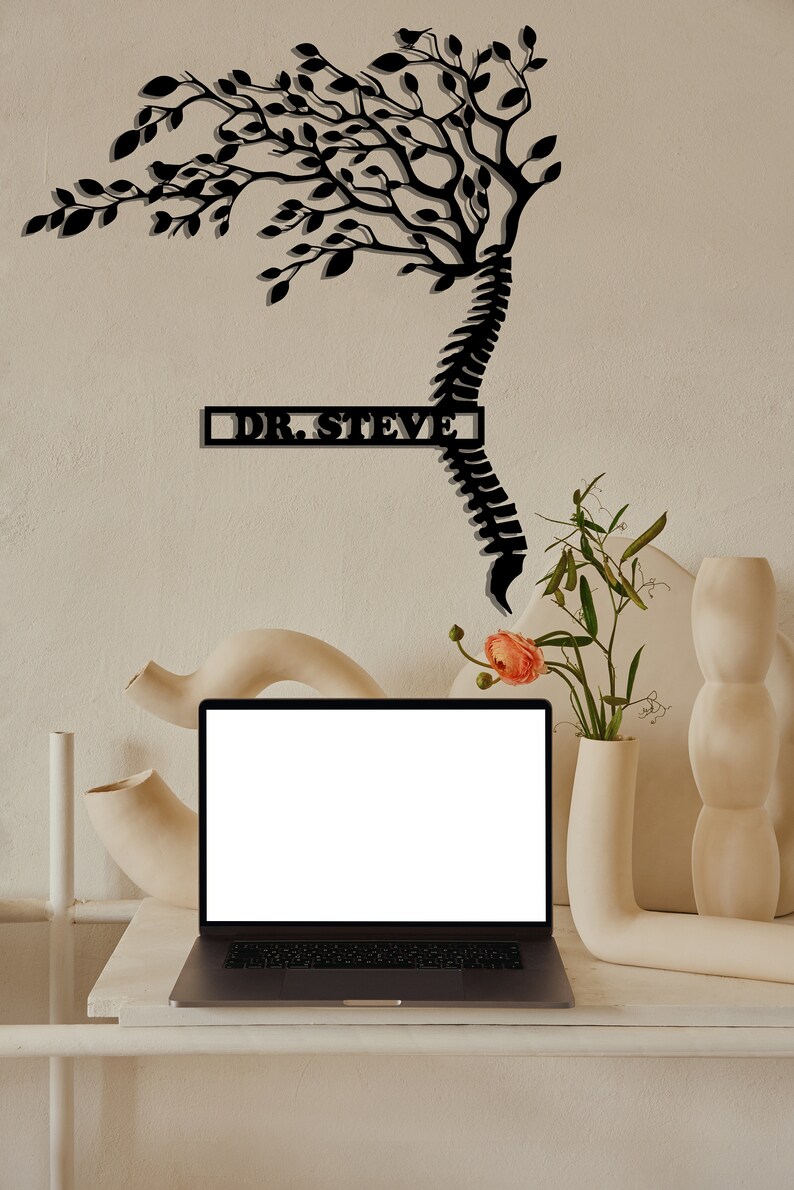 May include: A black metal wall art sculpture of a tree growing from a spine with the text "DR. STEVE" on a white wall. A laptop computer is on a white table in front of the wall art. There are white ceramic vases and flowers on the table.