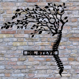 May include: Black metal wall art featuring a stylized tree with branches growing from a human spine. The tree is silhouetted against a brick wall. The text "DR. STEVE" is displayed on a rectangular plaque below the tree.
