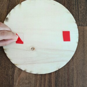 May include: A close-up of a round wooden surface with two pieces of red double-sided tape attached. The text "PRODUCTS WILL BE SHIPPED WITH DOUBLE-SIDED TAPE" is visible below the surface.