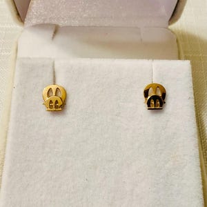 May include: A pair of gold-coloured stud earrings, each shaped like a stylised pig face. The earrings are displayed on a white, textured surface inside a light-coloured jewellery box. The earrings are small and delicate.