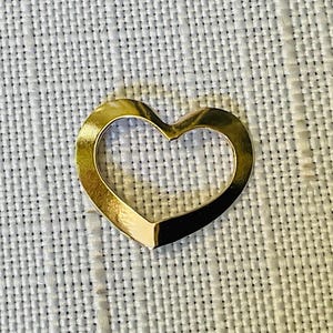 Peut inclure: Un pendentif en forme de cœur en métal doré avec un centre évidé et une surface lisse et réfléchissante. Le cœur présente un design angulaire distinct avec une base pointue et un sommet incurvé, créant une silhouette ouverte et épurée.