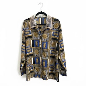 May include: A patterned long-sleeved shirt in blue, gold, black, and white, hanging on a black hanger. The shirt features a geometric design. The collar has a decorative trim. The background is plain white.