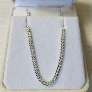 May include: A silver-coloured metal chain necklace featuring a classic curb link design. The chain is composed of uniform, interlocking oval links that lie flat against the surface, creating a consistent, polished, and reflective metallic appearance.