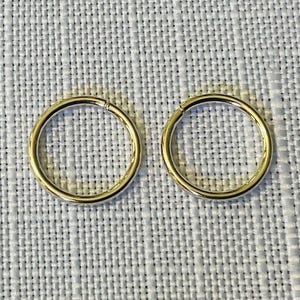 May include: Two gold-coloured, circular hoop earrings. The earrings are made of a shiny, metallic material and are set against a textured, light grey background. The hoops are simple in design and appear to be lightweight.